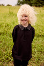 A smiling child standing in a grassy field, wearing a black long-sleeve polo shirt with a dark gray textured collar and black pants.