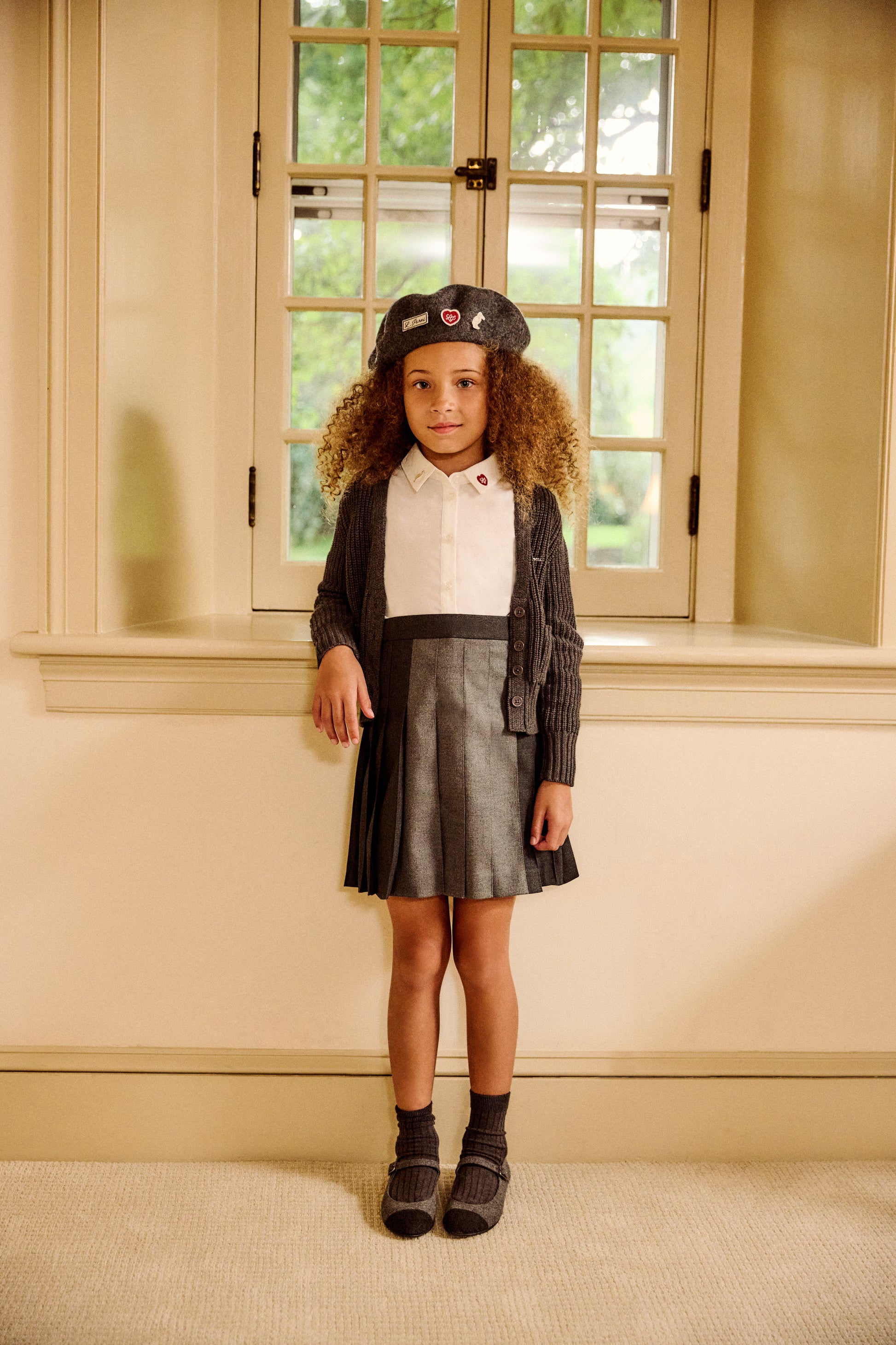 A young girl with curly brown hair stands indoors in front of a window wearing a dark gray and white dress with a matching dark gray cardigan, a dark gray beret, and dark socks.