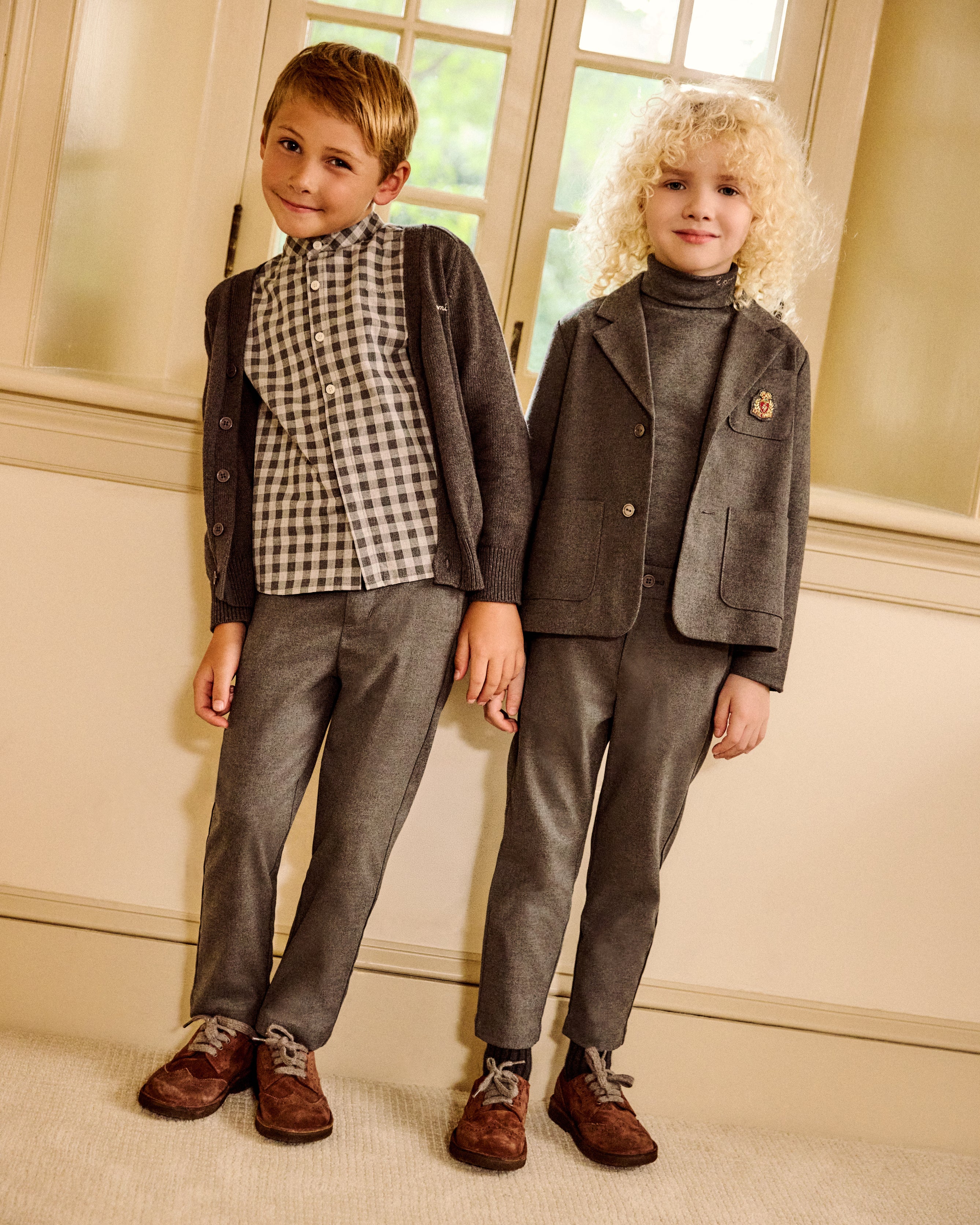 Two young boys modeling formal children's clothing in an indoor setting with cream walls and window light. One boy wears a brown checkered shirt with a brown blazer and gray trousers, and the other wears a gray turtleneck with a gray blazer and gray trousers.