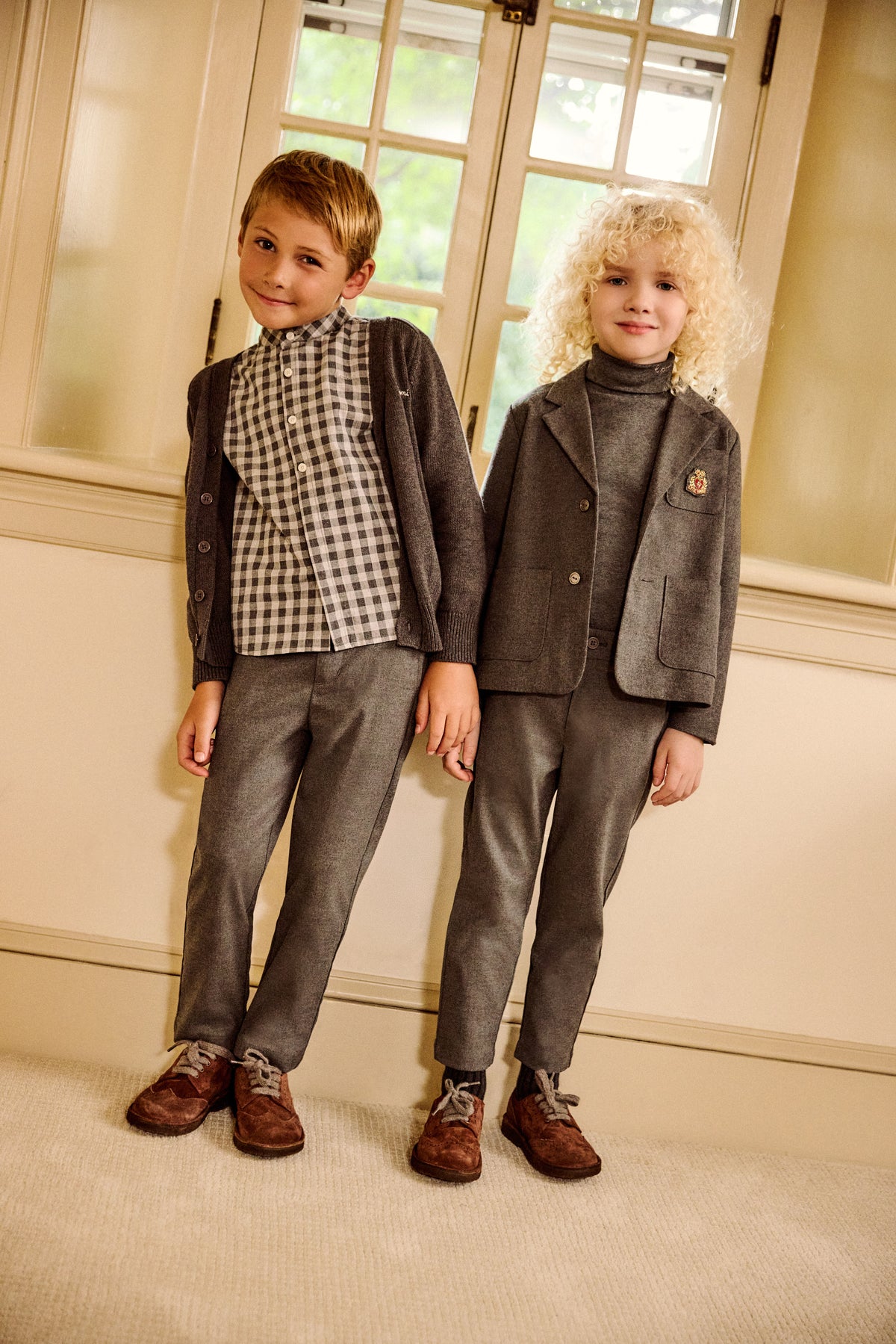 Two young boys modeling formal children's clothing in an indoor setting with cream walls and window light. One boy wears a brown checkered shirt with a brown blazer and gray trousers, and the other wears a gray turtleneck with a gray blazer and gray trousers.