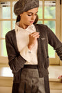 A woman wearing a brown ribbed cardigan, cream blouse, pleated gray skirt, and brown beret, standing in front of a softly lit window.