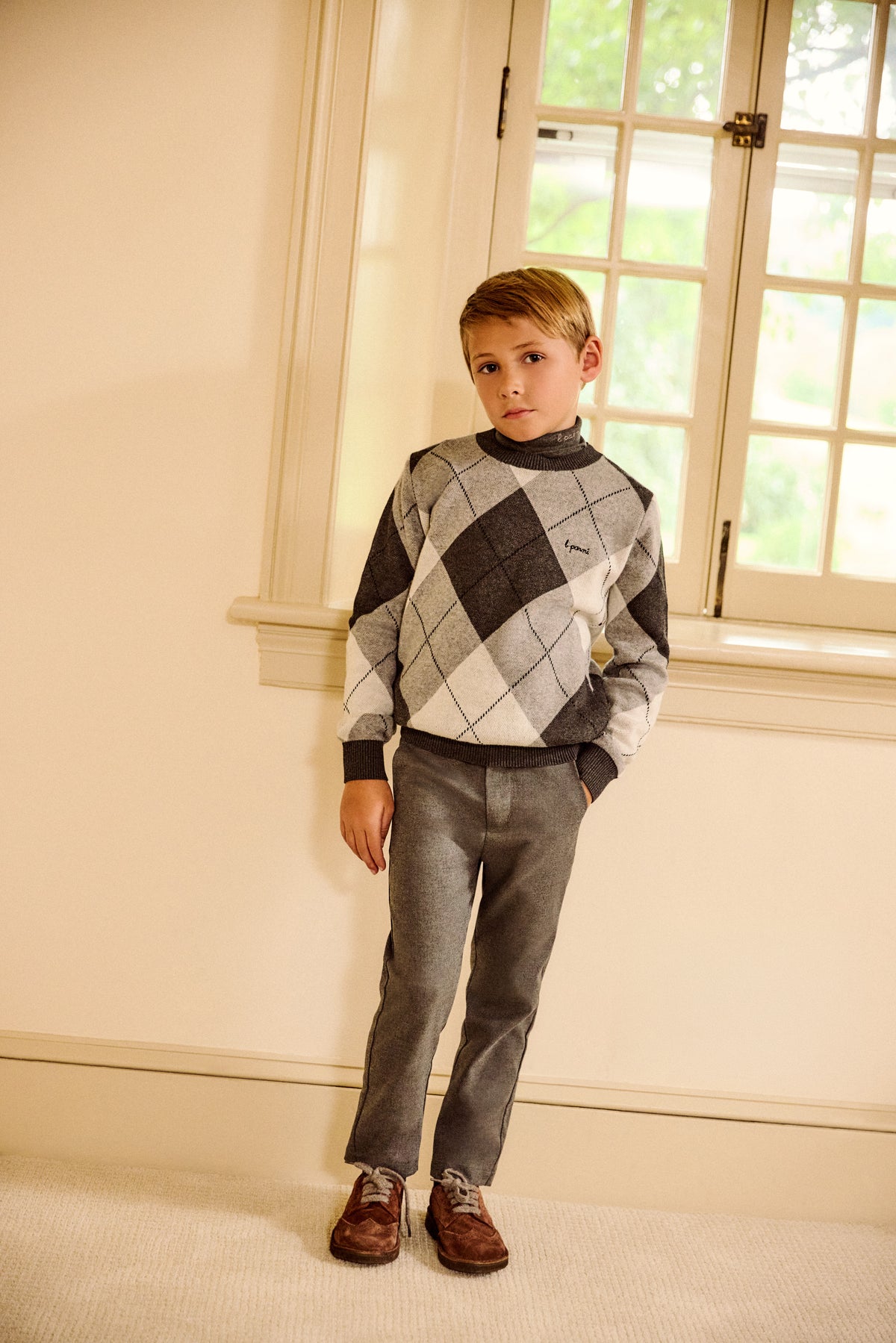 A young boy stands in a room wearing a gray and black argyle sweater, gray trousers, and brown shoes.