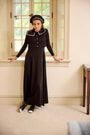 A teen wearing a black long-sleeve maxi dress with a white collar, matching black beret, and black-and-white shoes, leaning on a windowsill in a beige room.