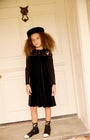 A young girl with curly brown hair stands in front of a white paneled door wearing a black long-sleeve dress with a decorative collar, a black beret, and black-and-white shoes.