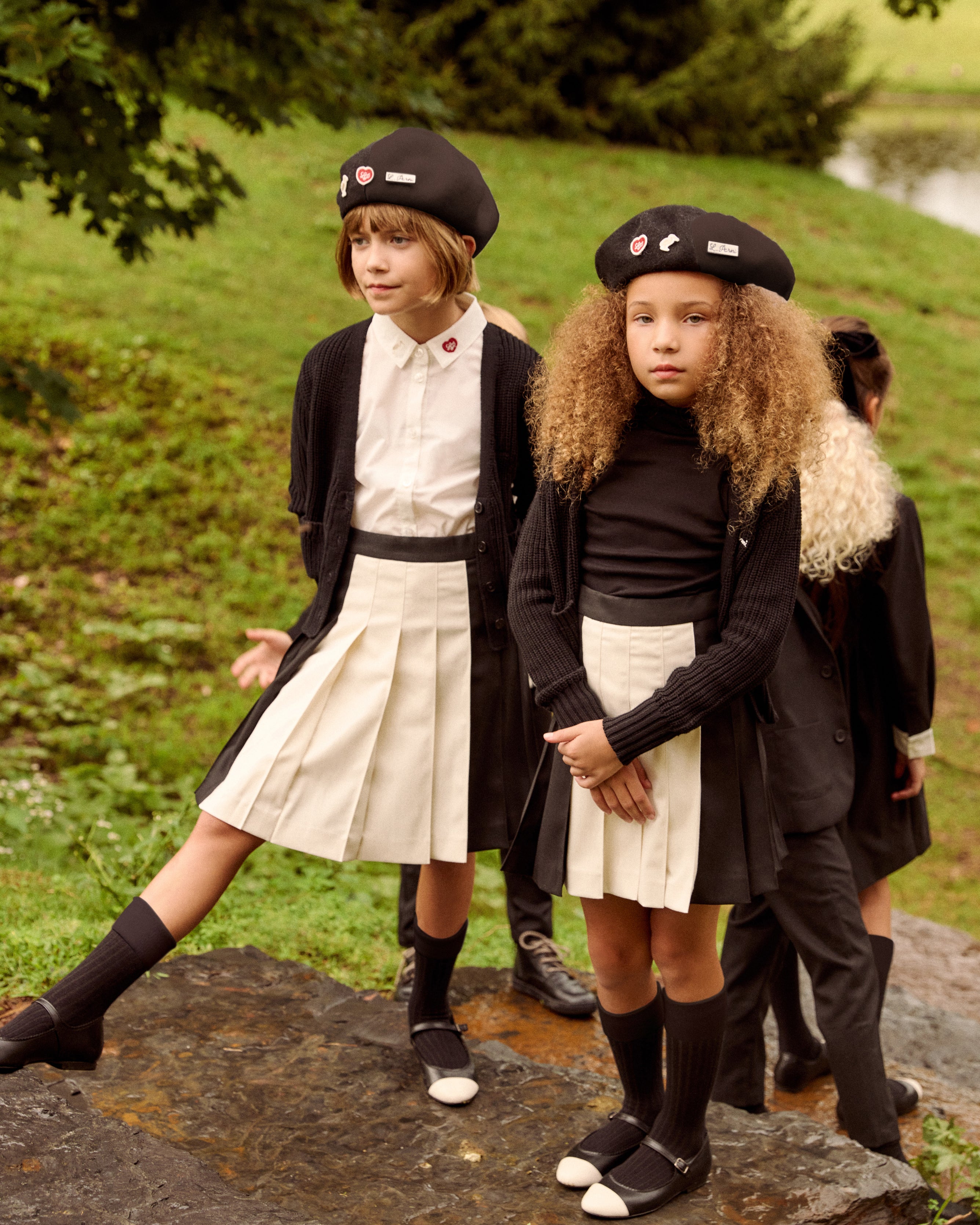 Two young girls wearing matching school uniform outfits with black berets, black cardigans, white collars, pleated cream skirts, and black knee-high socks, standing outdoors on a mossy rock in a grassy park setting.