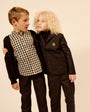 Two children modeling formal clothing against a plain beige studio background. The boy wears a black cardigan over a black-and-white gingham shirt with black pants, and the girl wears a black blazer and matching pants over a black turtleneck.