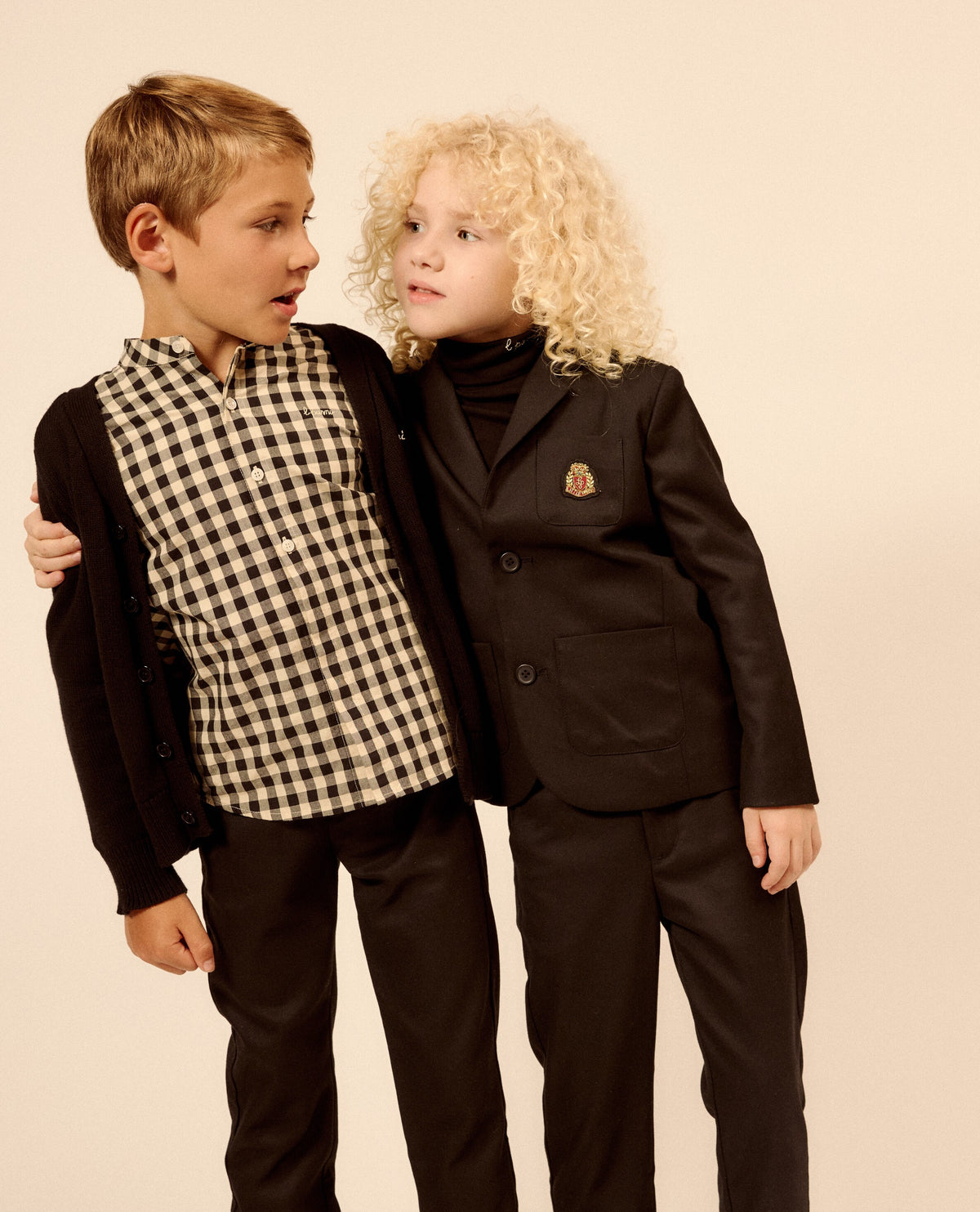 Two children modeling formal clothing against a plain beige studio background. The boy wears a black cardigan over a black-and-white gingham shirt with black pants, and the girl wears a black blazer and matching pants over a black turtleneck.