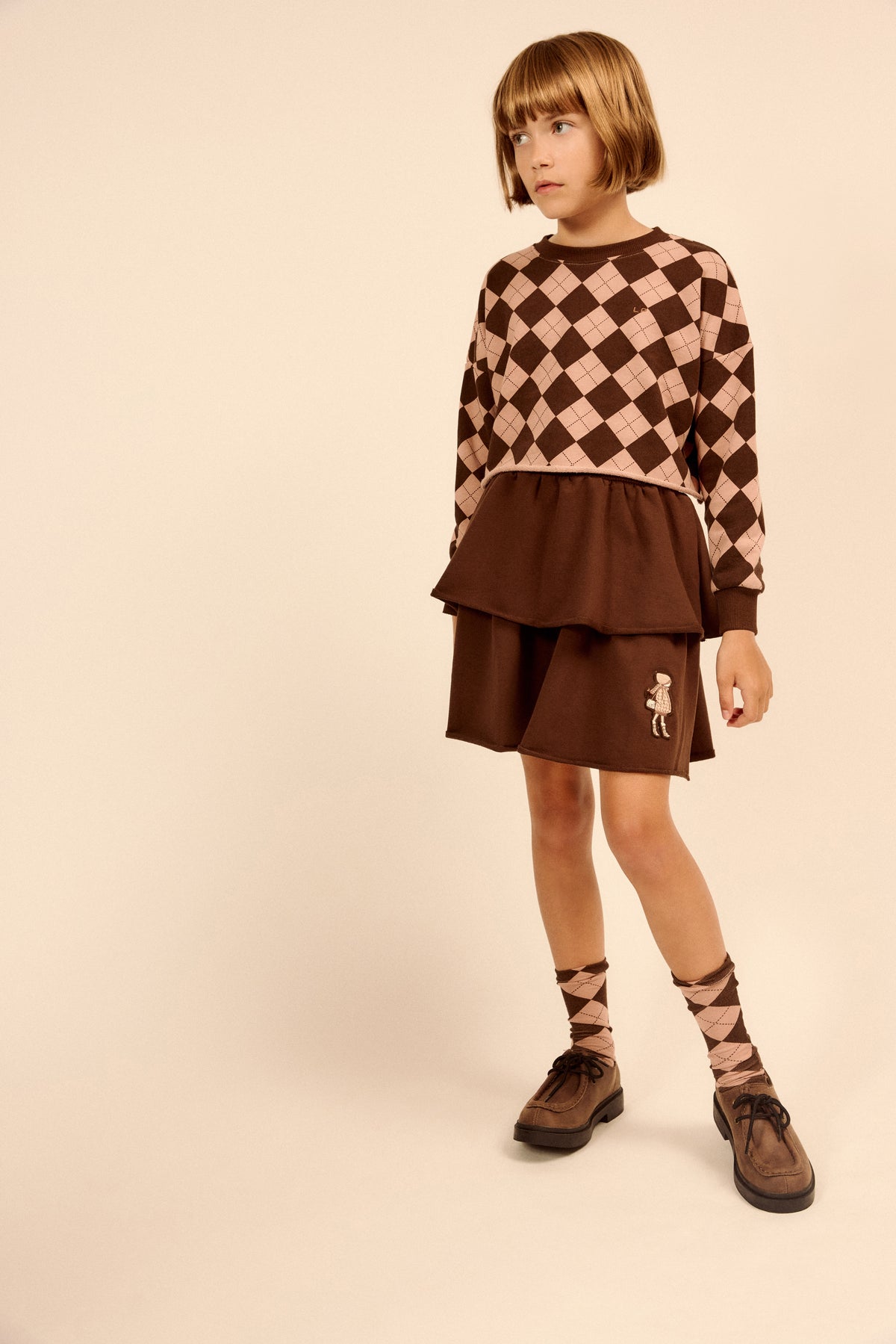 A young girl modeling a brown and beige argyle sweater and matching brown skirt set, paired with patterned socks and brown shoes, against a solid beige studio background.