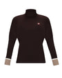 A dark brown long-sleeve turtleneck top with beige ribbed cuffs and a small red and white logo patch on the left chest, displayed on a clean white background.