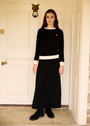 A woman stands in front of a white paneled door wearing a black long-sleeve sweater and matching black maxi skirt with contrasting white trim, black ankle boots, and a small red embroidered heart on the chest.