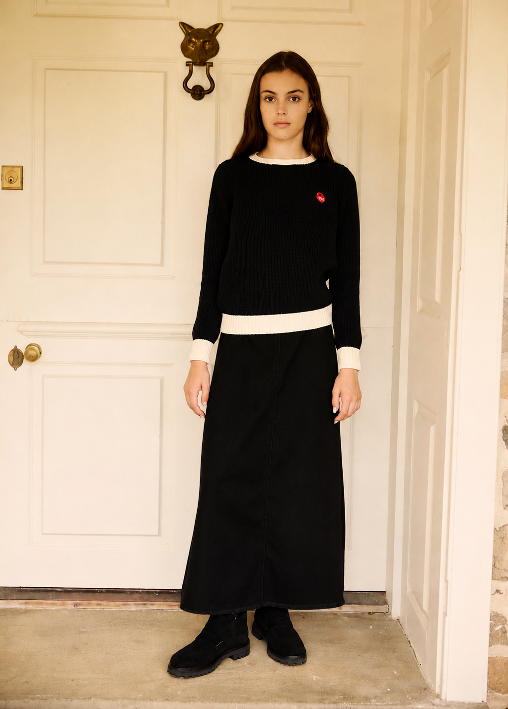 A woman stands in front of a white paneled door wearing a black long-sleeve sweater and matching black maxi skirt with contrasting white trim, black ankle boots, and a small red embroidered heart on the chest.