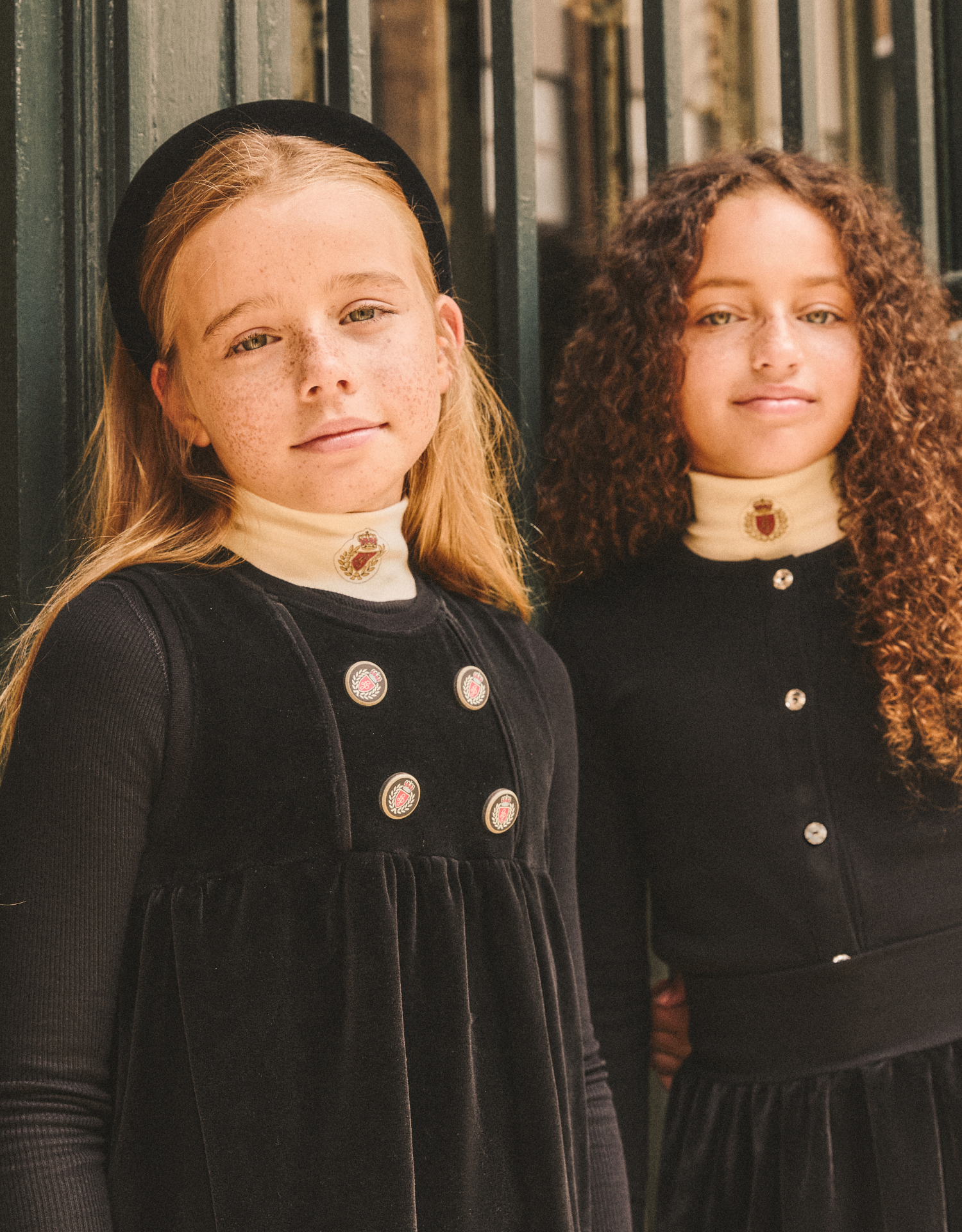 Two girls wearing black velour sleeveless dresses with double-breasted tops and monochrome buttons.