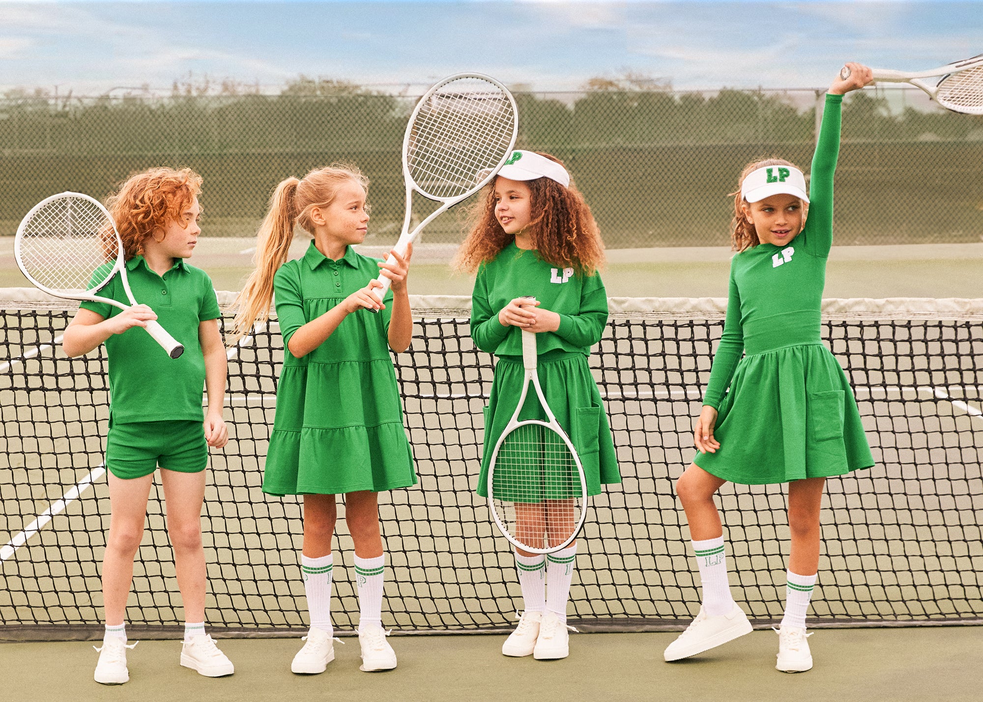 4 children standing outside on a tennis court wearing matching green shorts, tops, skirts and dress