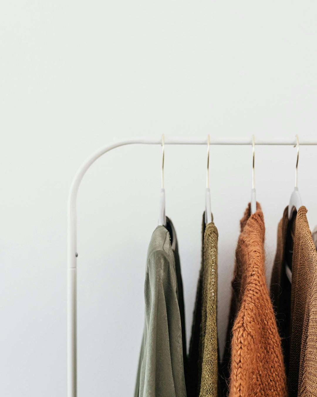 A white metal clothing rack with several garments hanging on hangers against a plain white wall. The visible garments include muted green, olive, rust orange, and brown textured fabrics.