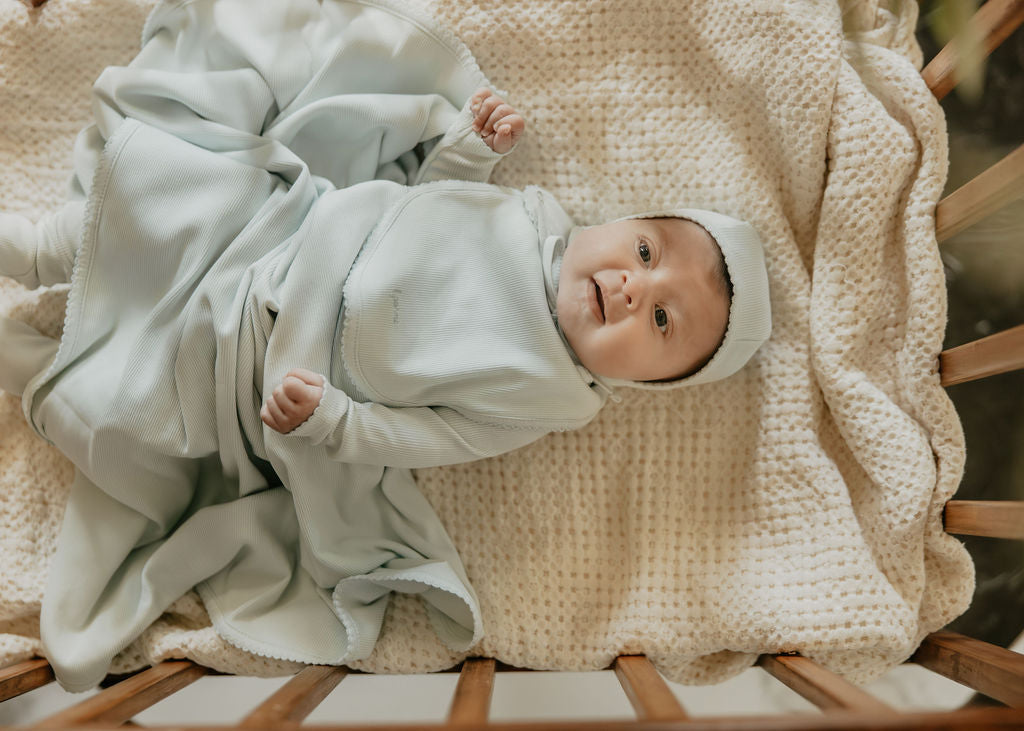 Newborn baby wrapped in a light blue blanket on a wooden crib with a textured beige blanket.
