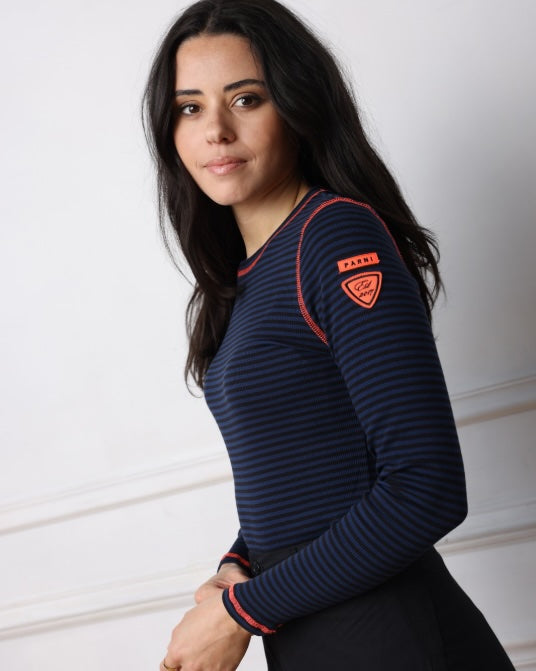 A woman wearing a navy blue and black horizontally striped long-sleeve athletic top with red piping and a red Parni brand patch on the upper sleeve, posed against a white paneled wall.